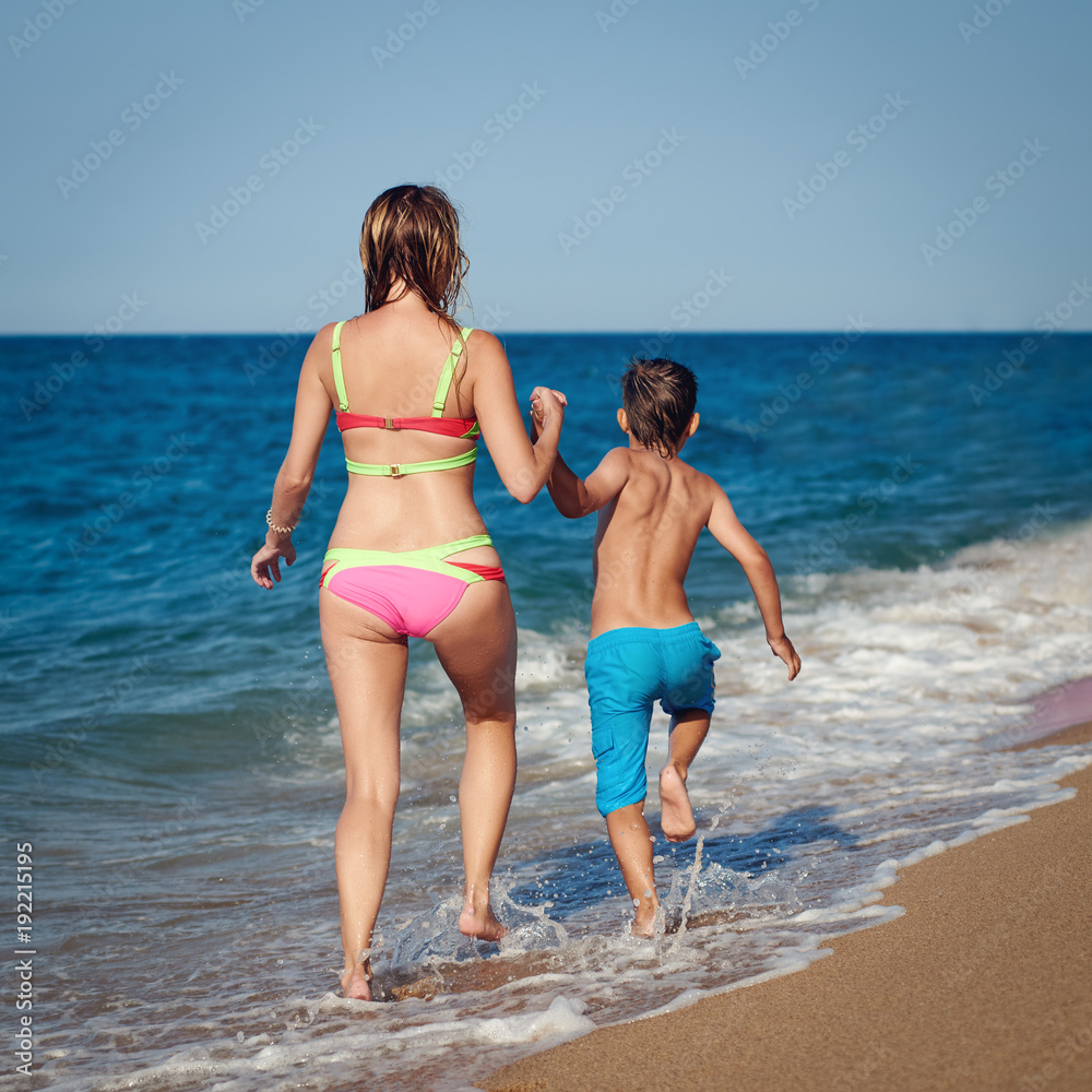 Mom and son are running along the sea shore. They are holding hands and moving away from the camera.