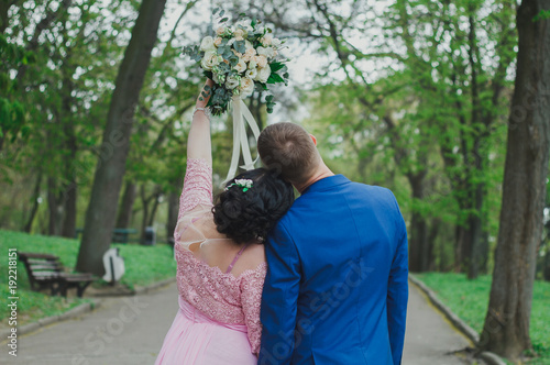 Wedding couple in green park. Groom's and bride's heads neat each other. Curvy woman in pink lace evening dress. Wedding bouquet with white, beige roses and eustoma with eucalyptus. Plus size bride