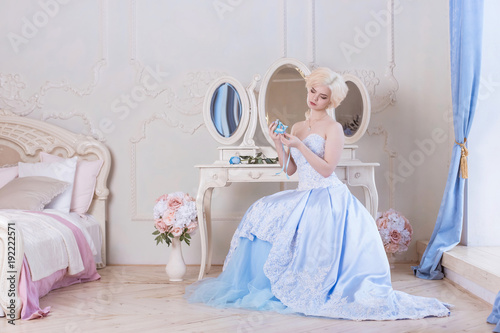 A beautiful, young blonde woman sits by the dressing table and holds a blue mask dressed in a ball gown, going to the ball. Princess in the Versailles, France.