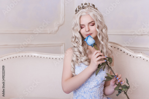 A glamorous girl, a blond woman in a crown holds a blue rose and sniffs it. Aroma of flowers, Cinderella, Princess in the castle. A rich queen in a dress. Place your text here. photo