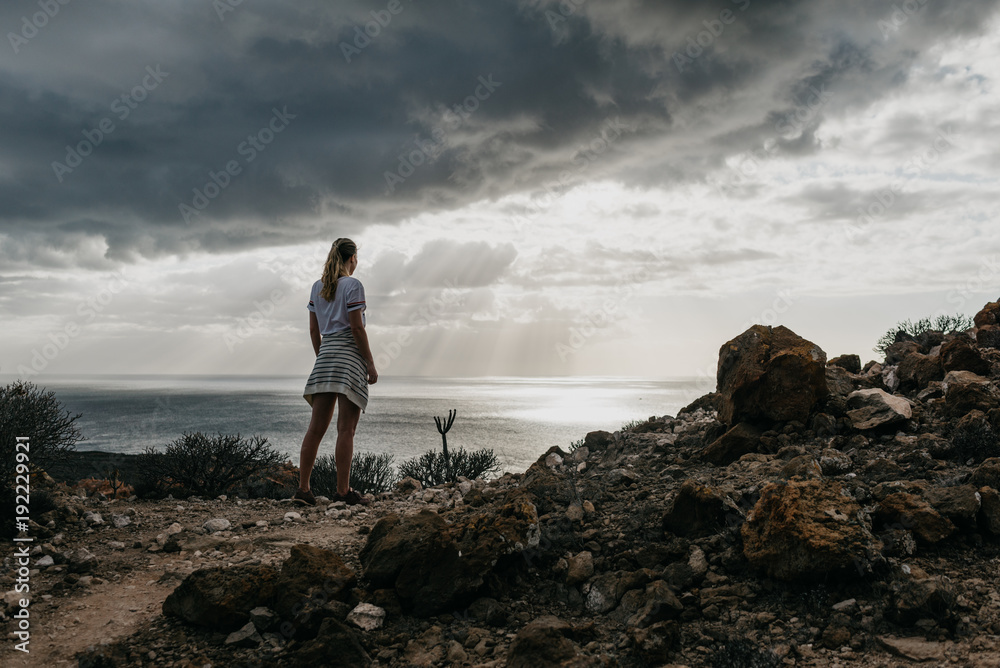 The beautiful girl stands and watches the ocean on the top of the rocks in the evening. Tenerife