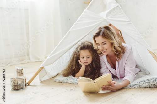 Photo happy mother and daughter reading book together in kid wigwam