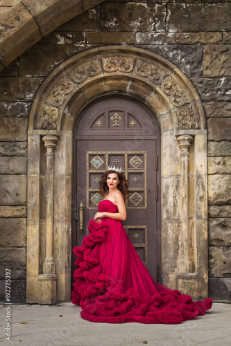 A beautiful woman, a queen in a red luxurious dress, stands against the backdrop of a medieval, medieval, gothic door with an arch. Medieval fantasy. Attractive Princess © Надежда Манахова