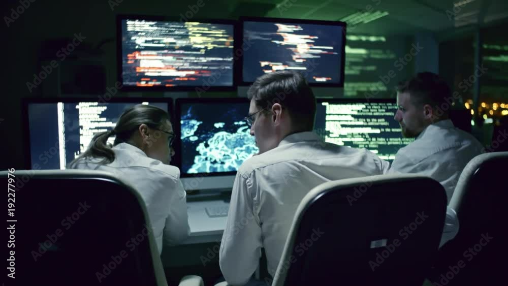 Rear view of three cyber security specialists talking at desk in front of multi-display workstation in dark IT office at night