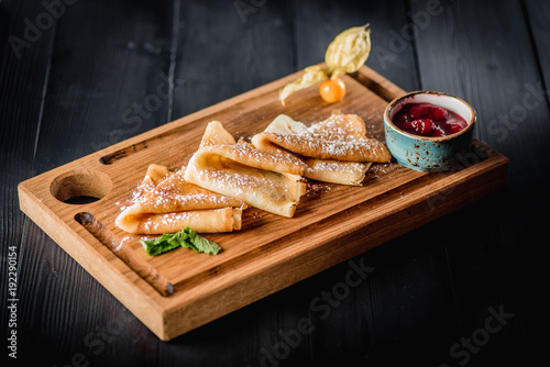 Pancakes with sugar powder and jam on a wooden board. Dark table, empty space. Maslenitsa, russian carnival