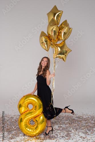 Sexy, beautiful girl in black dress is holding big golden balloon. Star-shaped balloons. Confetti.