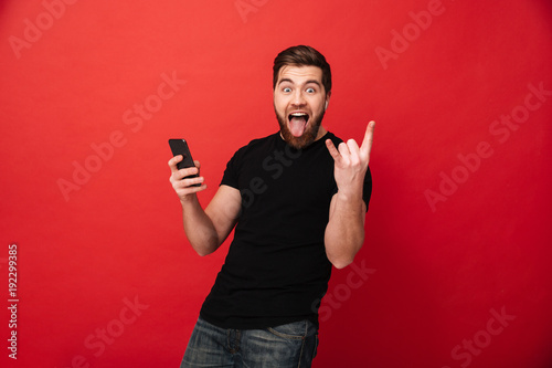Photo of crazy happy guy screaming in fascination while holding mobile phone in hand and showing rock sign, isolated over red background