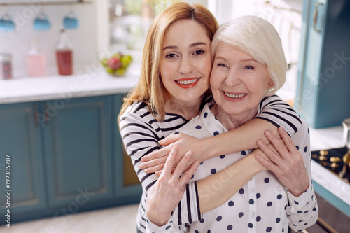 Daughterly love. Cheerful young woman hugging her elderly mother and smiling at the camera happily together with her while standing in the kitchen