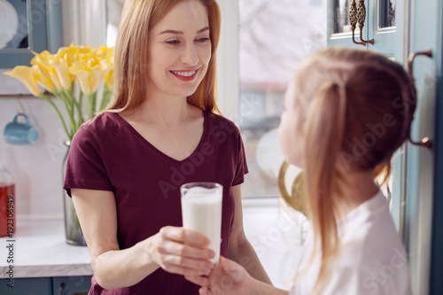 Taste it. Charming young woman standing in the kitchen near her daughter and giving her a glass of milk while smiling at her fondly