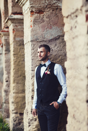 Groom at wedding tuxedo smiling and waiting for bride near brick buildingl. Elegant groom in costume and bow-tie.