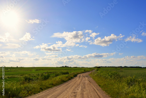  picturesque countryside landscape: a sandy rural road amidst green fields against a blue sky with a bright sun and clouds