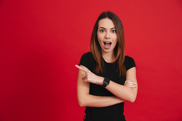 Excited optimistic woman with open mouth in black t-shirt looking at camera and pointing index finger aside on copyspace, isolated over red background