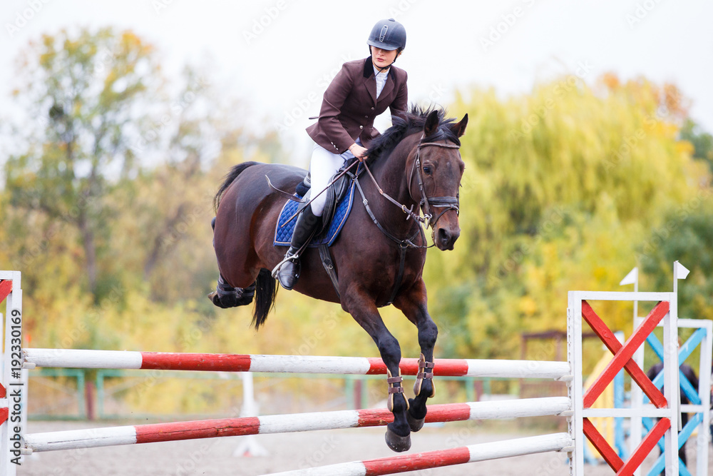Φωτογραφία Young rider girl on bay horse jumping over barrier on equestrian sport competition