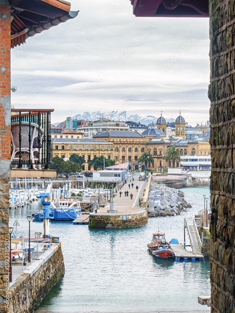 Fototapeta premium San Sebastian (Donostia) pejzaż miejski w pochmurny zimowy dzień z ośnieżonymi górami na horyzoncie.