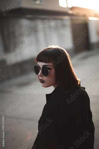 A beautiful young woman with black glasses is posing on the street at sunset