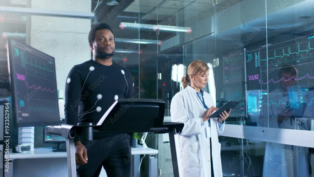 Male Athlete Walks on a Treadmill with Electrodes Attached to His Body while Sport Scientist Supervises Reads EKG Status. In the Background Laboratory with High-Tech Equipment. Male Athlete Walks on a Treadmill with Electrodes Attached to His Body while Sport Scientist Supervises Reads EKG Status. In the Background Laboratory with High-Tech Equipment.