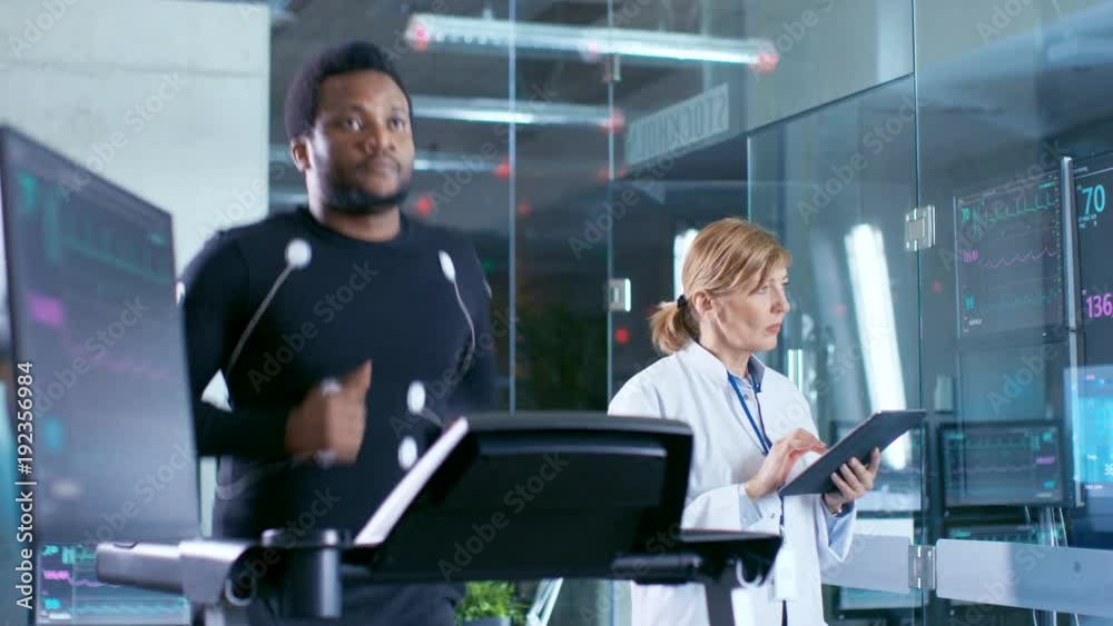 Male Athlete Runs on a Treadmill with Electrodes Attached to His Body while Sport Scientist Holds Tablet and Supervises EKG Status. In the Background Laboratory with High-Tech Equipment. Male Athlete Runs on a Treadmill with Electrodes Attached to His Body while Sport Scientist Holds Tablet and Supervises EKG Status. In the Background Laboratory with High-Tech Equipment.