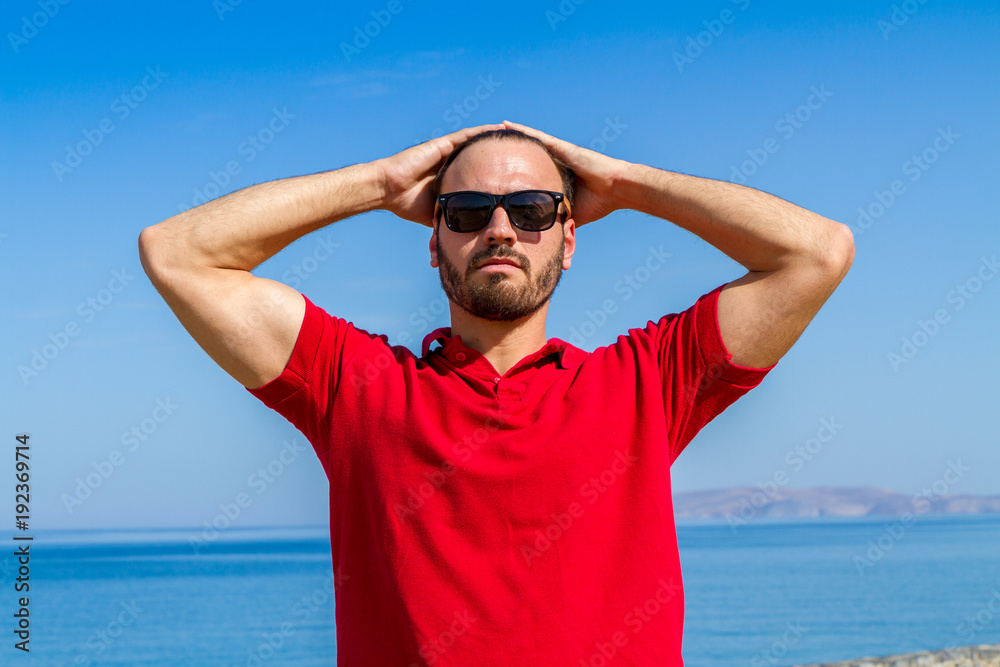 Carefree confident bearded young in red t-shirt sunglasses in summer blue sky background. Carefree confident bearded young in red t-shirt sunglasses in summer blue sky background.