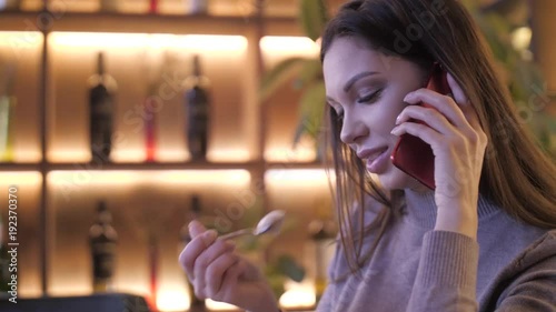 Slow motion. Woman in cafe with big cup of coffee, cappuccino, latte. Eat a milk foam using a spoon. Cheerful beautiful young woman having break, sitting in cafe with cup of cappuccino, use smartphone photo