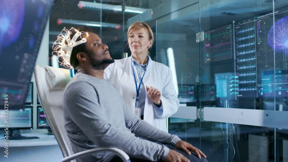 Man Wearing Brainwave Scanning Headset Sits in a Chair while Scientist Adjusts the Device. In the Modern Brain Study Laboratory Monitors Show EEG Reading and Brain Model. 