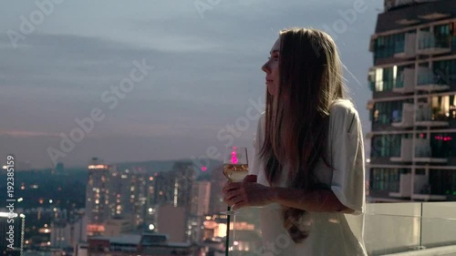 Happy woman drinking a glass of white wine while sitting in rooft bar in the city or on the terrace with cityscape at sunset. Vacation,happiness,freedom concept. photo