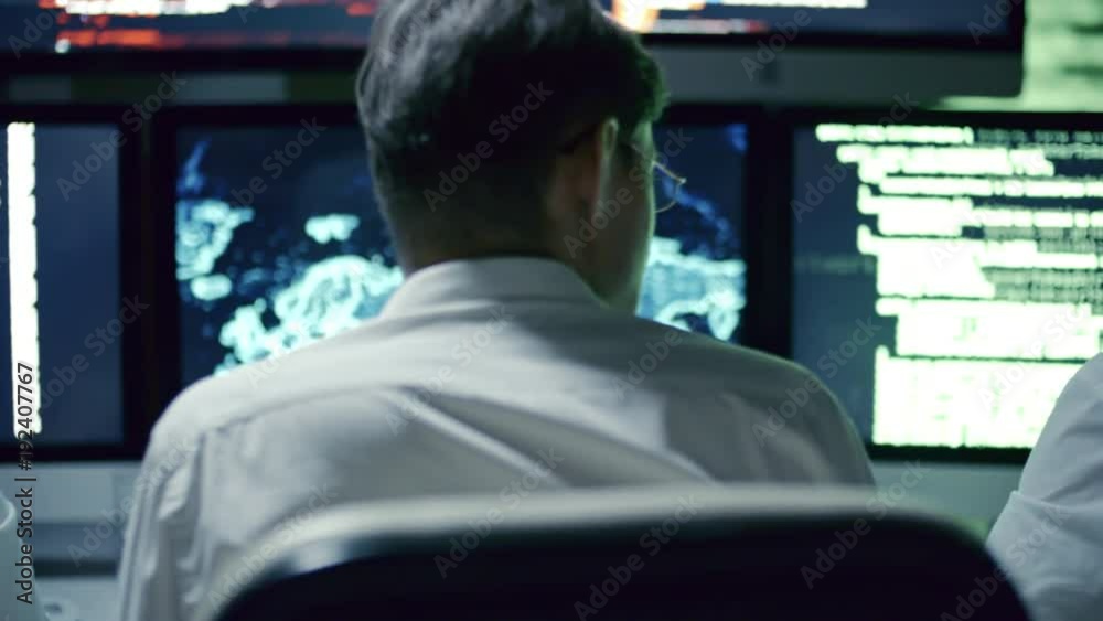 Rear view of professional team of IT security specialists talking in front of multi-display workstation in dark office at night; computer code and holographic world map on display