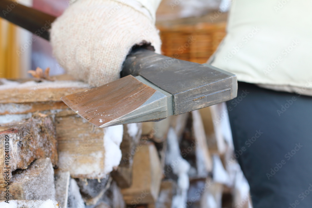 The axe for chopping wood in a woman's hand on the background of a stack of birch firewood on the terrace of a country house in winter.