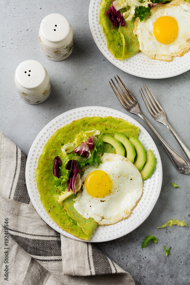 Spinach green crepes (pancakes) with fried egg, avocado and leaves of mix of salad on ceramic plate on gray concrete background. Сoncept of healthy breakfast. Selective focus. Top view. Copt space. Spinach green crepes (pancakes) with fried egg, avocado and leaves of mix of salad on ceramic plate on gray concrete background. Сoncept of healthy breakfast. Selective focus. Top view. Copt space.