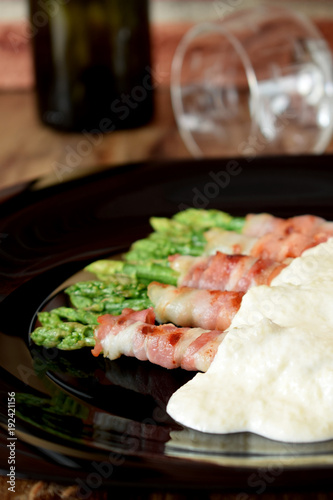 Green asparagus wrapped into smoked bacon covered with white sauce on a black plate