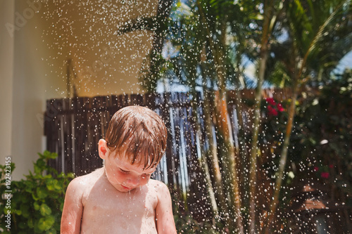 Cute little boy with blonde hair standing under the jets of water. Hot sunny day. Green tropical plants in the background.