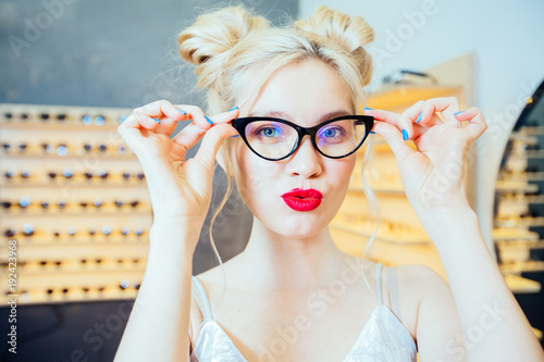 Portrait of fashion woman with red lips and funny hairstyle wearing and buying sunglasses with optician modern shop interior behind her. Close up. photo