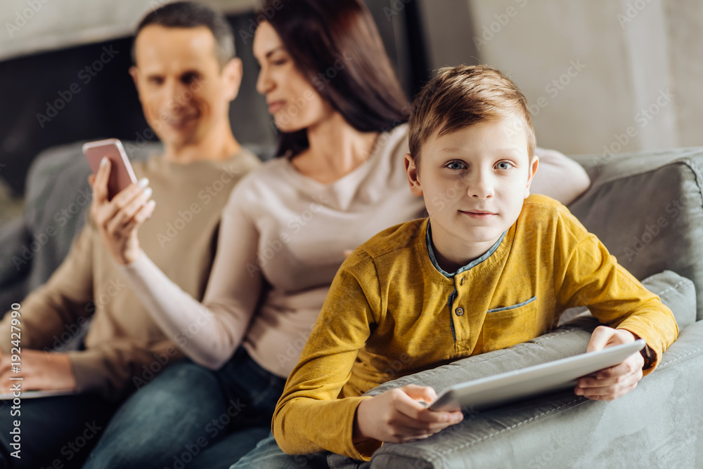 On his own. Adorable pre-teen boy leaning on the sofa armrest and posing for the camera, playing on his tablet while his parents watching something on the phone On his own. Adorable pre-teen boy leaning on the sofa armrest and posing for the camera, playing on his tablet while his parents watching something on the phone