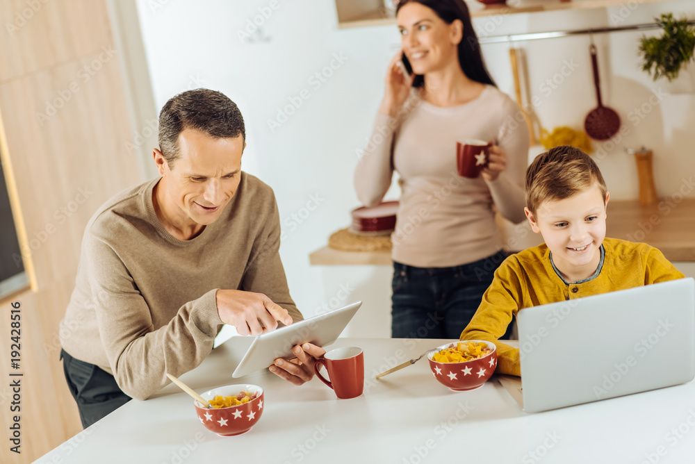Different priorities. Pleasant young family using their devices instead of eating their breakfast, the woman talking on the phone and father and son playing on their gadgets Different priorities. Pleasant young family using their devices instead of eating their breakfast, the woman talking on the phone and father and son playing on their gadgets