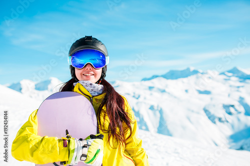 Photo of smiling brunette in helmet and mask with snowboard on background of snowy hills