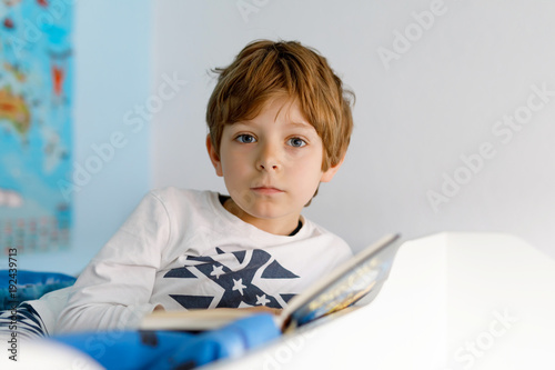 Cute blond little kid boy in pajamas reading book in his bedroom