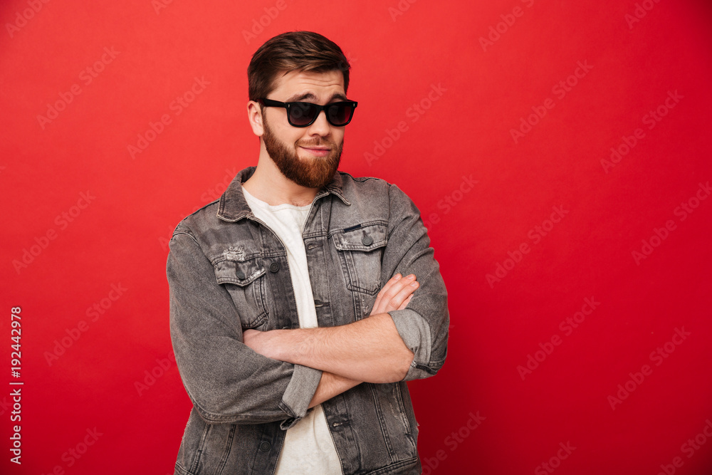Plakat Portret nieodparty młody facet jest ubranym okulary przeciwsłonecznych i drelichowy ono uśmiecha się i pozuje z rękami krzyżować, odizolowywający nad czerwonym tłem