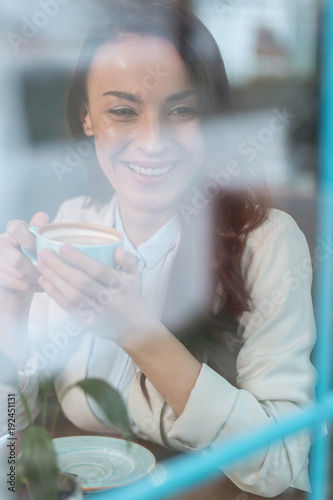 Positive vibes. Gay merry young woman taking cup while gazing aside and sitting at the table