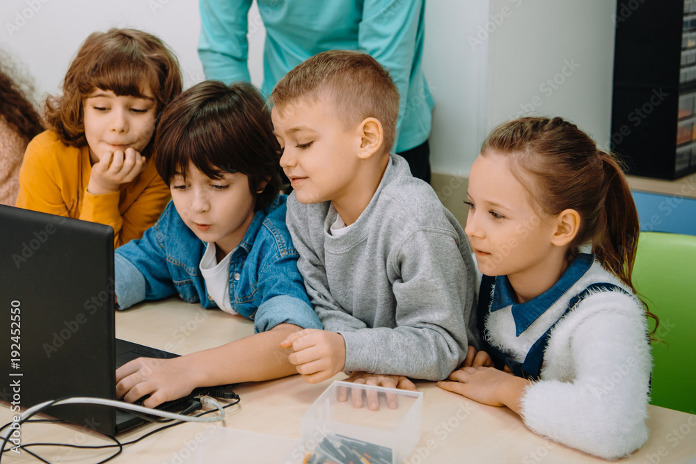 group of kids working with laptop together while teacher standing behind them group of kids working with laptop together while teacher standing behind them