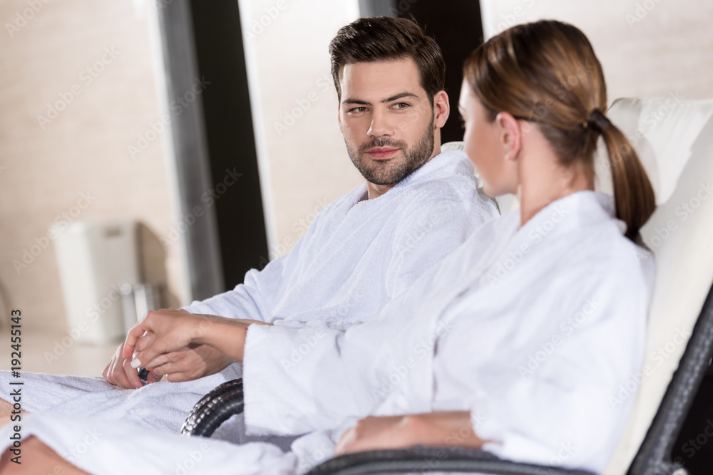 young couple in bathrobes holding hands and looking at each other while resting together in spa center young couple in bathrobes holding hands and looking at each other while resting together in spa center