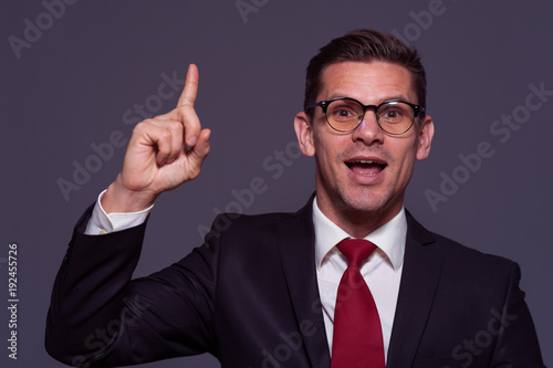 Portrait in violet tone of young positive businessman wearing glasses and formal suit pointing his finger up against gray background