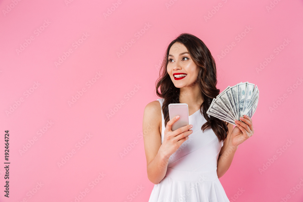 Cheerful asian woman in dress holding fan of money in dollar cash and smartphone while looking aside on copyspace, isolated over pink background Cheerful asian woman in dress holding fan of money in dollar cash and smartphone while looking aside on copyspace, isolated over pink background