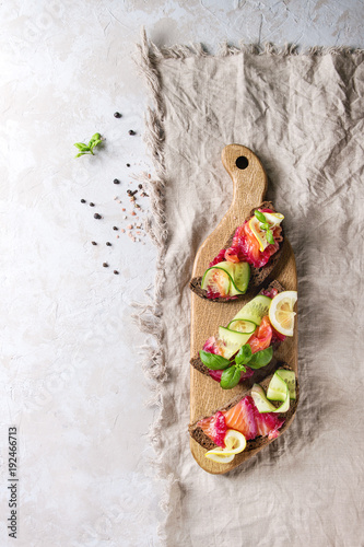 Sliced beetroot marinated salmon sandwiches with rye bread, cucumber, basil and lemon served on wooden cutting board over grey texture background with textile linen. Top view, space photo