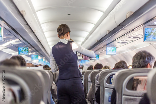 Interior of commercial airplane with flight attandant serving passengers on seats during flight. Stewardess in dark blue uniform walking the aisle. Horizontal composition. photo