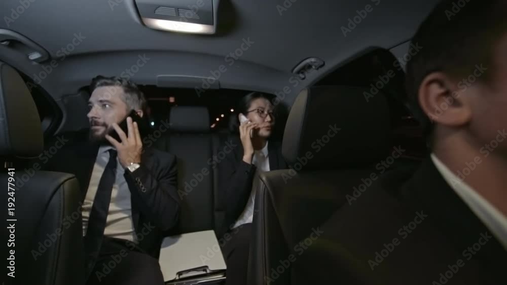 Medium shot of bearded businessman in suit and Asian businesswoman sitting in backseat of moving car and talking on mobile phones