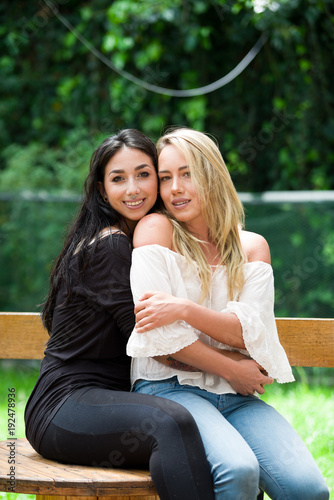 A pair of proud lesbian in outdoors sitting on a wooden table, brunette woman is hugging a blonde woman, in a garden background photo