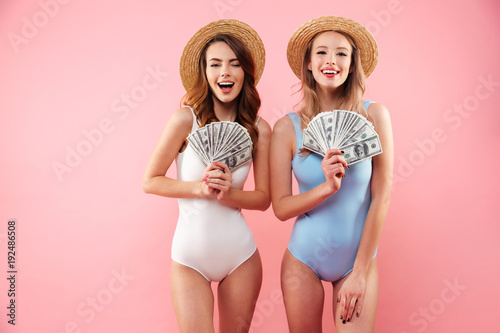 Two successful women on vacation in summer clothing rejoicing and holding fans of money dollar banknotes in hands, isolated over pink background