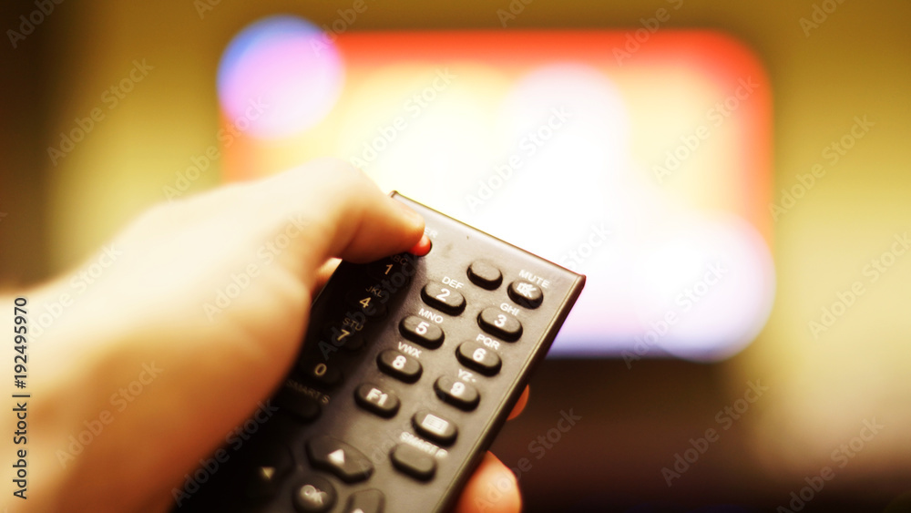 Obraz premium Black remote control in the hand against blurred background of turned on TV on the wall with bokeh effect. Finger is on red POWER button, selective focus