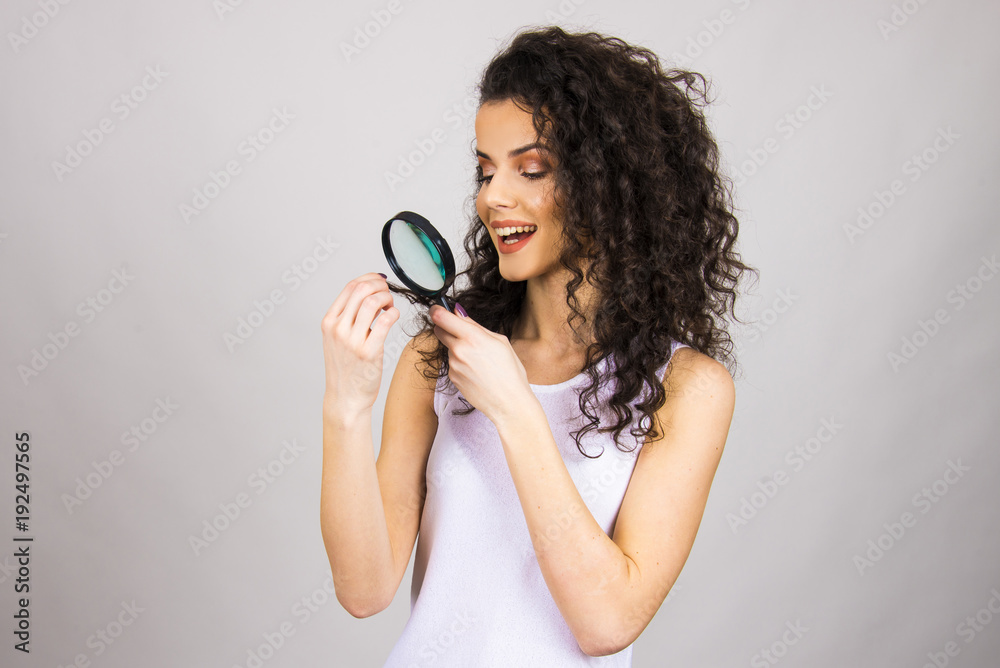 Obraz premium Beautiful girl with long curly hair and magnifier looking close up on her hair top analysing damage and dry hair concept