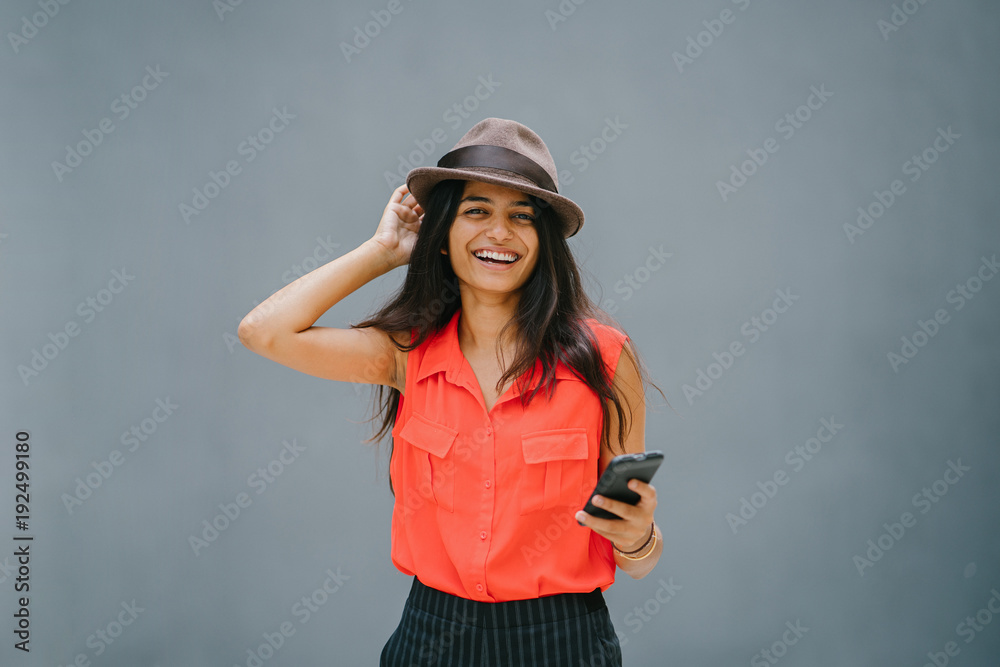 Fototapeta premium A picture of a young and attractive Asian Indian woman standing on a plain gray background. While using a smartphone, she smiles and wears a casual outfit.