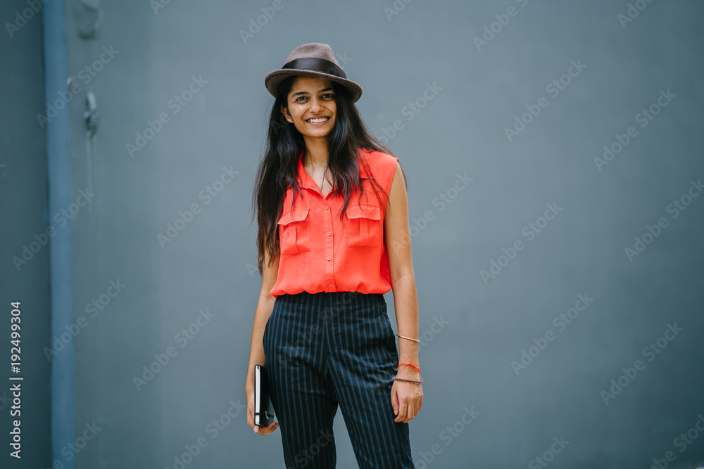 Portrait of a fashionable Indian Asian woman against a plain grey background. She is smiling and wearing a smart orange blouse and black pinstripe pants with a brown fedora hat. Portrait of a fashionable Indian Asian woman against a plain grey background. She is smiling and wearing a smart orange blouse and black pinstripe pants with a brown fedora hat.